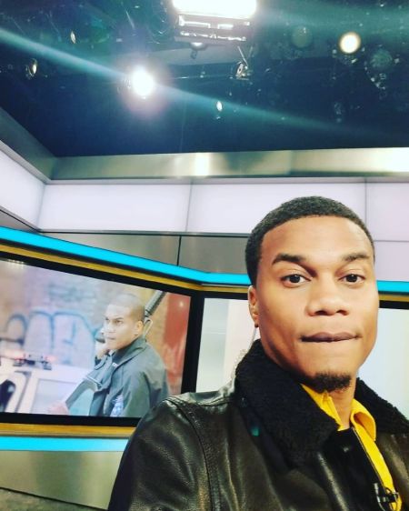 Cory Hardrict in a black leather jacket poses in front of a tv screen.